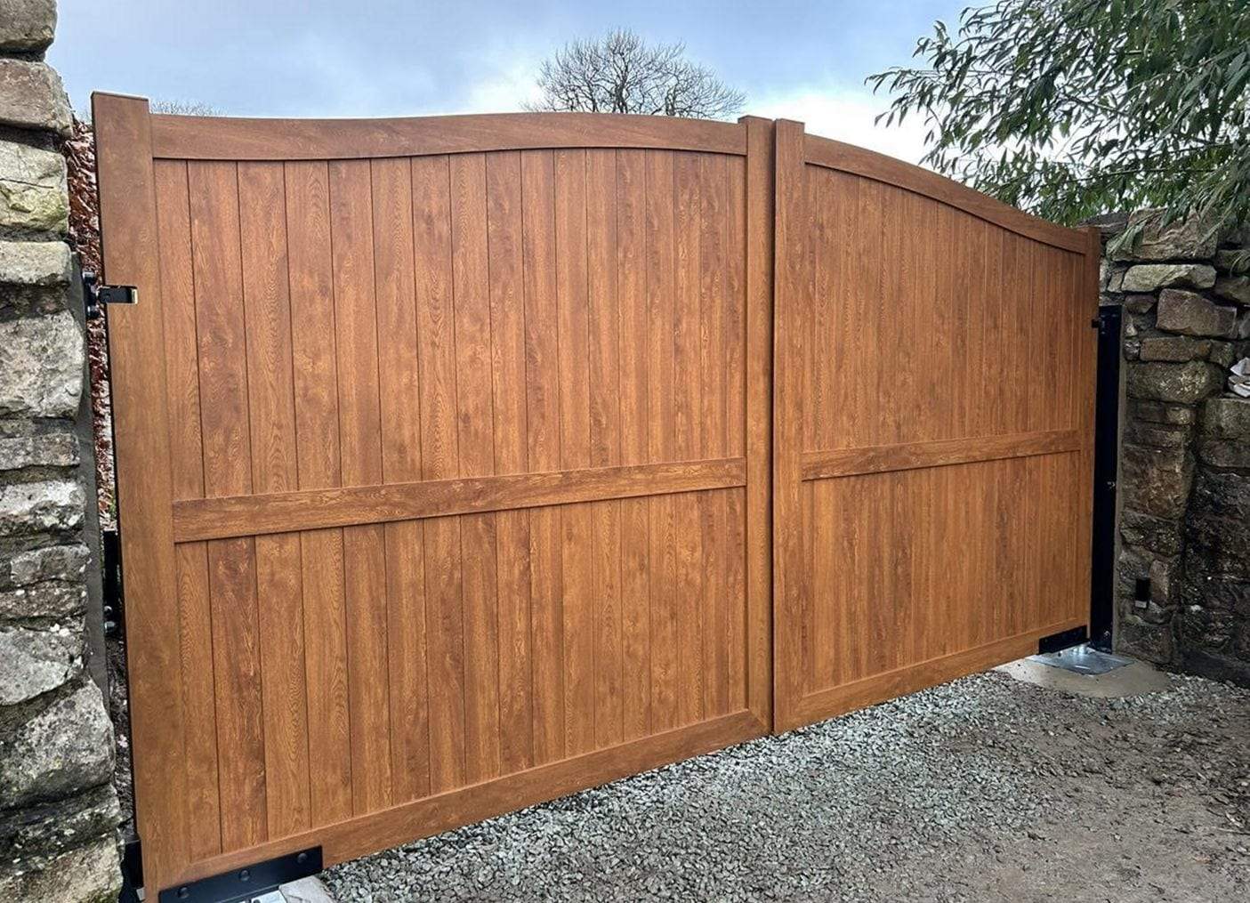 Pair of woodgrain-finish swing gates with an arched top rail and a horizontal mid rail, closed between dry-stone walls across a gravel drive, with black hinge brackets and ground plates at the posts