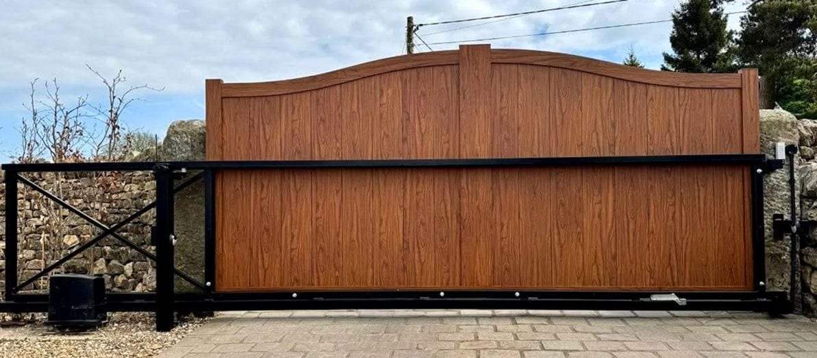 Woodgrain-finish slide gate with a curved top mounted on a black steel cantilever frame with cross bracing, a ground-mounted operator at the left end, closed across a block-paved drive in front of a stone wall