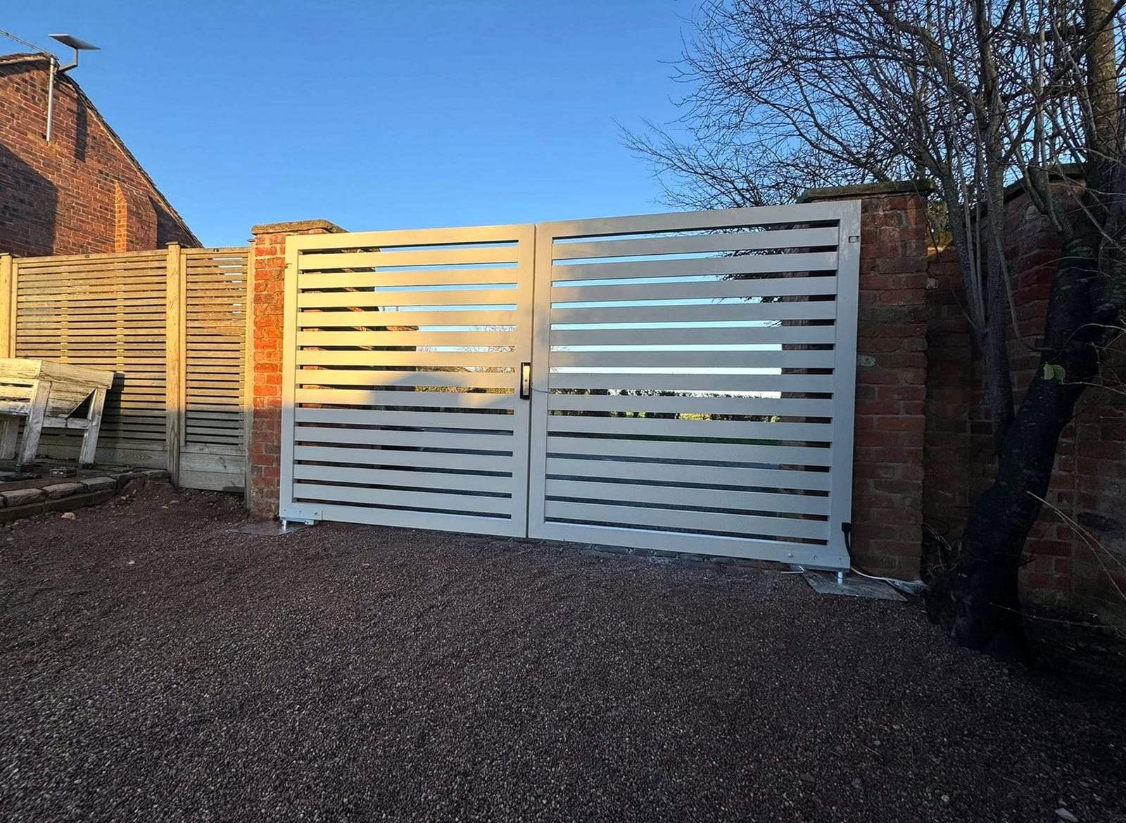 Pale gray double swing gates made of wide horizontal slats with narrow gaps between them, hung between red brick pillars across a gravel drive, with a handle and lock where the two leaves meet