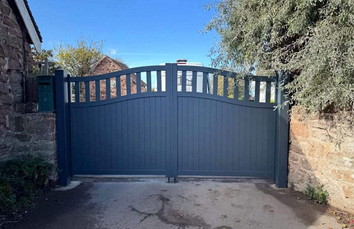 Slate-blue double swing gates with solid lower panels and an open band of slats under an arched top rail, hung between square posts beside an old stone wall, with a green mailbox on the left post