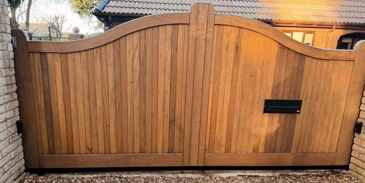 Natural hardwood double swing gates with tight vertical boards and an arched top frame, a black letter slot in the right leaf, hung between pale brick piers over a gravel drive
