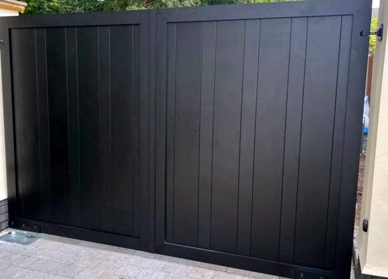Close view of black flat-top double swing gates with framed vertical board panels on a paver drive, with the steel cover plate of an underground operator showing at the bottom left hinge