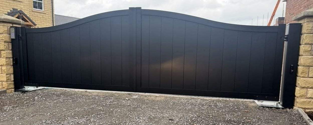 Wide black double swing gates with a sweeping arched top between sandstone pillars, a keypad and a photo-eye sensor on the post at the left pillar, steel operator plates at both hinges and a drainage channel along the gravel drive
