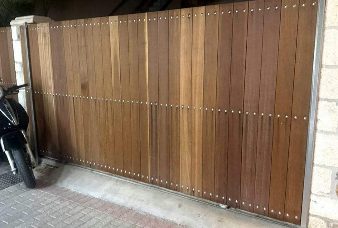 Slide gate of vertical hardwood boards fastened with rows of stainless studs inside a slim steel frame, closed on its ground track across a paved entrance beside a block wall, with a parked scooter at the left edge