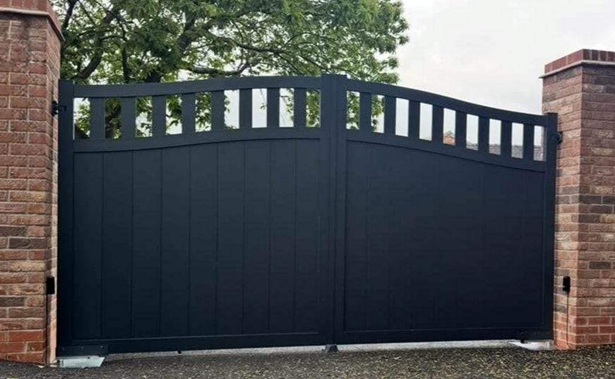 Black double swing gates with a slatted band under an arched top, hung between red brick piers, with a small black photo-eye safety sensor mounted on each pier