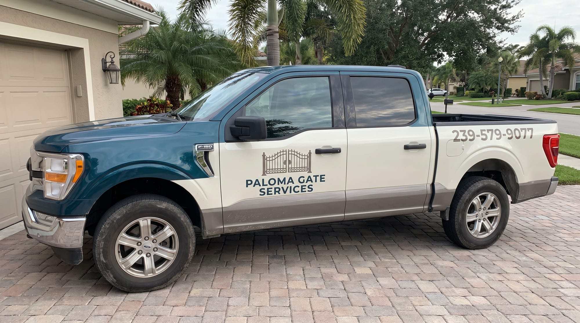 Paloma Gate Services pickup truck with a dark teal cab, off-white doors and bed and warm gray lower panels, the gate logo and company name on the door and 239-579-9077 on the bed, parked on a paver driveway with palm trees and Florida homes behind